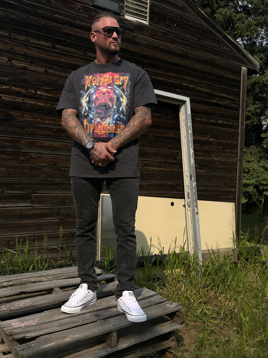 Male model wearing Wolves Cry Oversized Universe Tee in vintage black, styled with white sneakers and black jeans, standing on a wood pallet in front of a rustic backdrop.