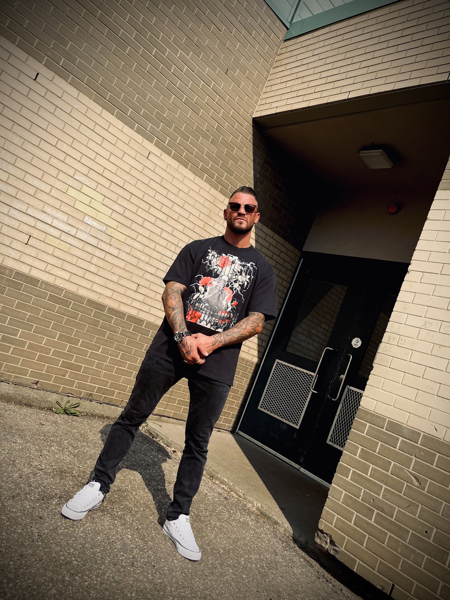 Full look in Wolves Cry Prayer Tee styled with black jeans and shades, standing against brick wall in urban light.