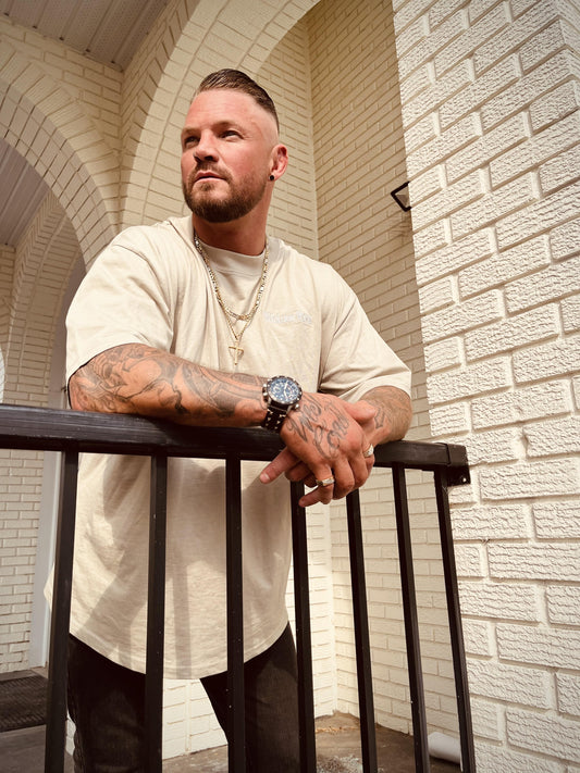 Model leaning on railing wearing the Wolves Cry organic high neck drop tee in sage dust, showcasing relaxed fit and embroidery.