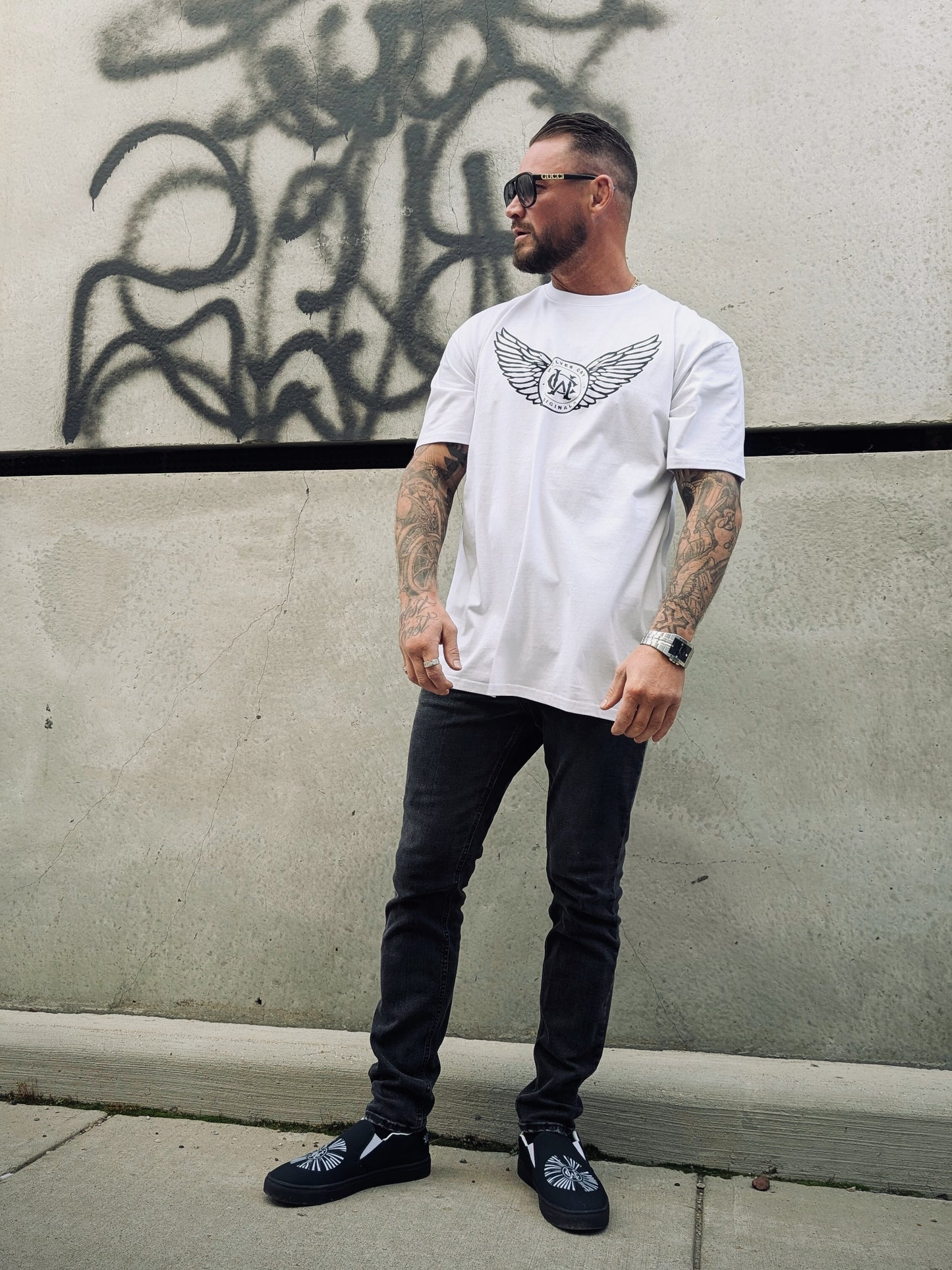 Model wearing white WC Flight Academy Tee with monogram wings, posed in front of graffiti wall.
