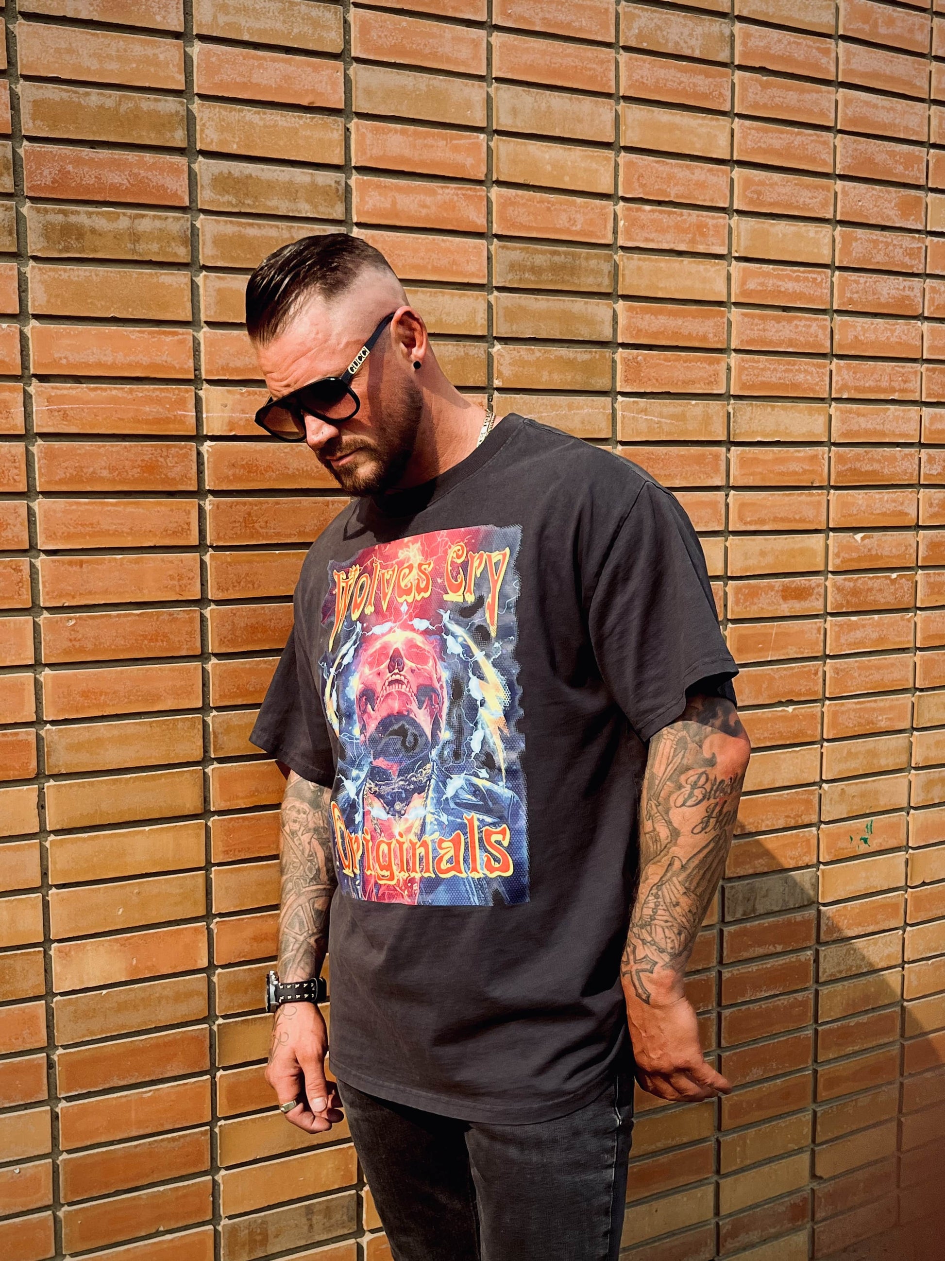 Side-angled view of male model in Wolves Cry Oversized Universe Tee, standing against a red-brick wall in full sunlight.
