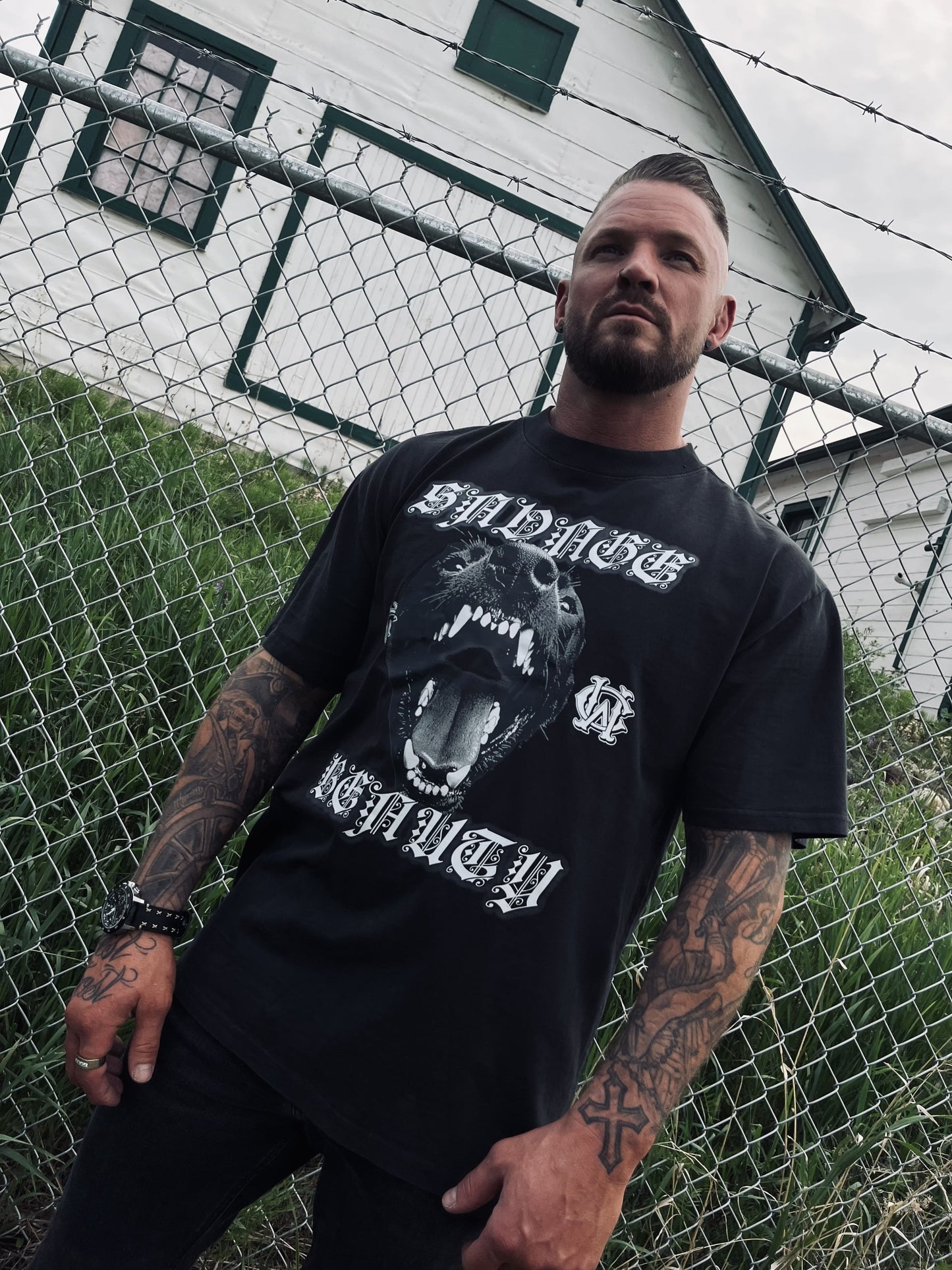 Male model wears Wolves Cry Savage Beauty Tee styled with all-black streetwear in front of chainlink fence