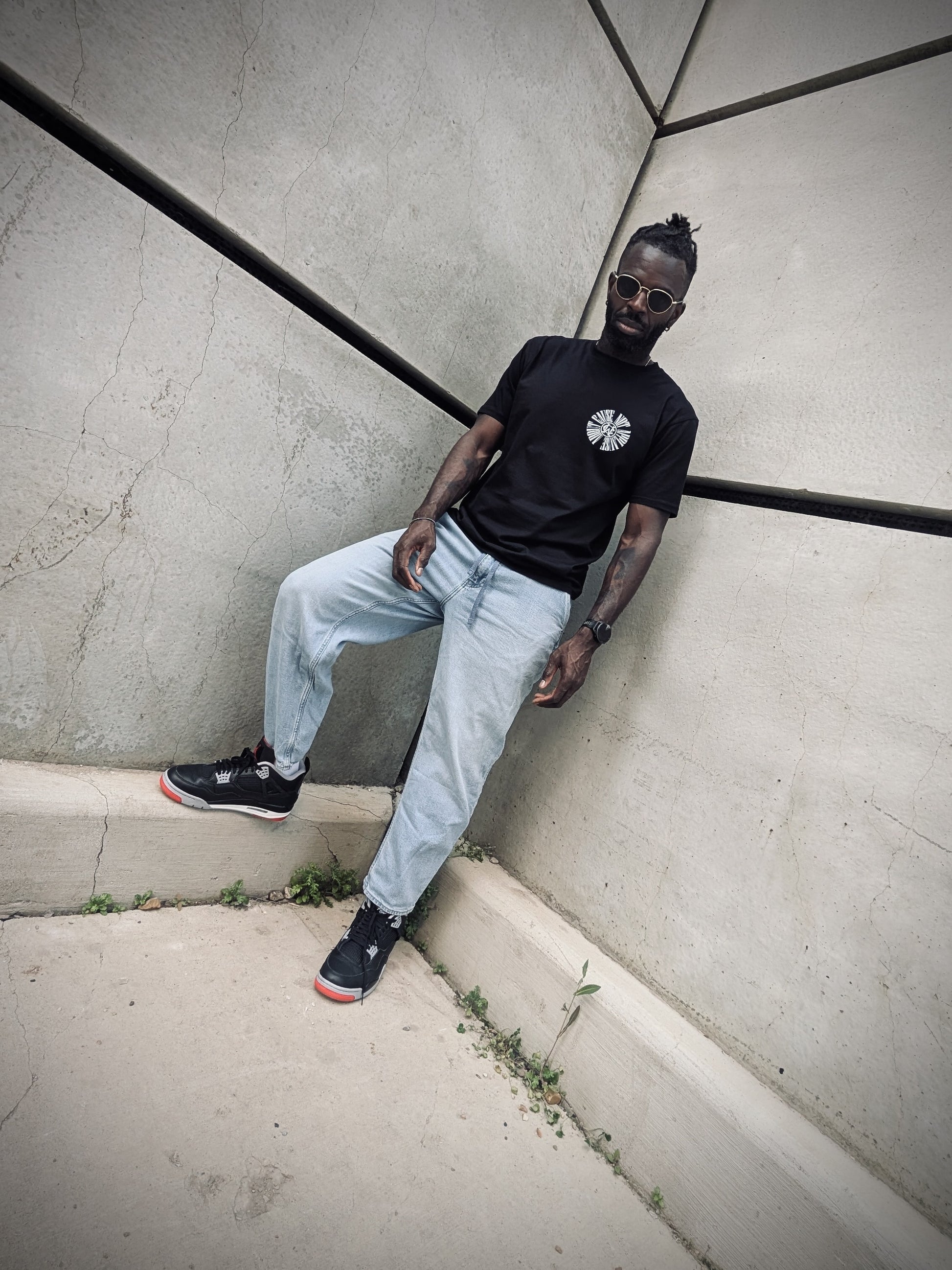 Full-body shot of model leaning against wall in black WC tee and light jeans.