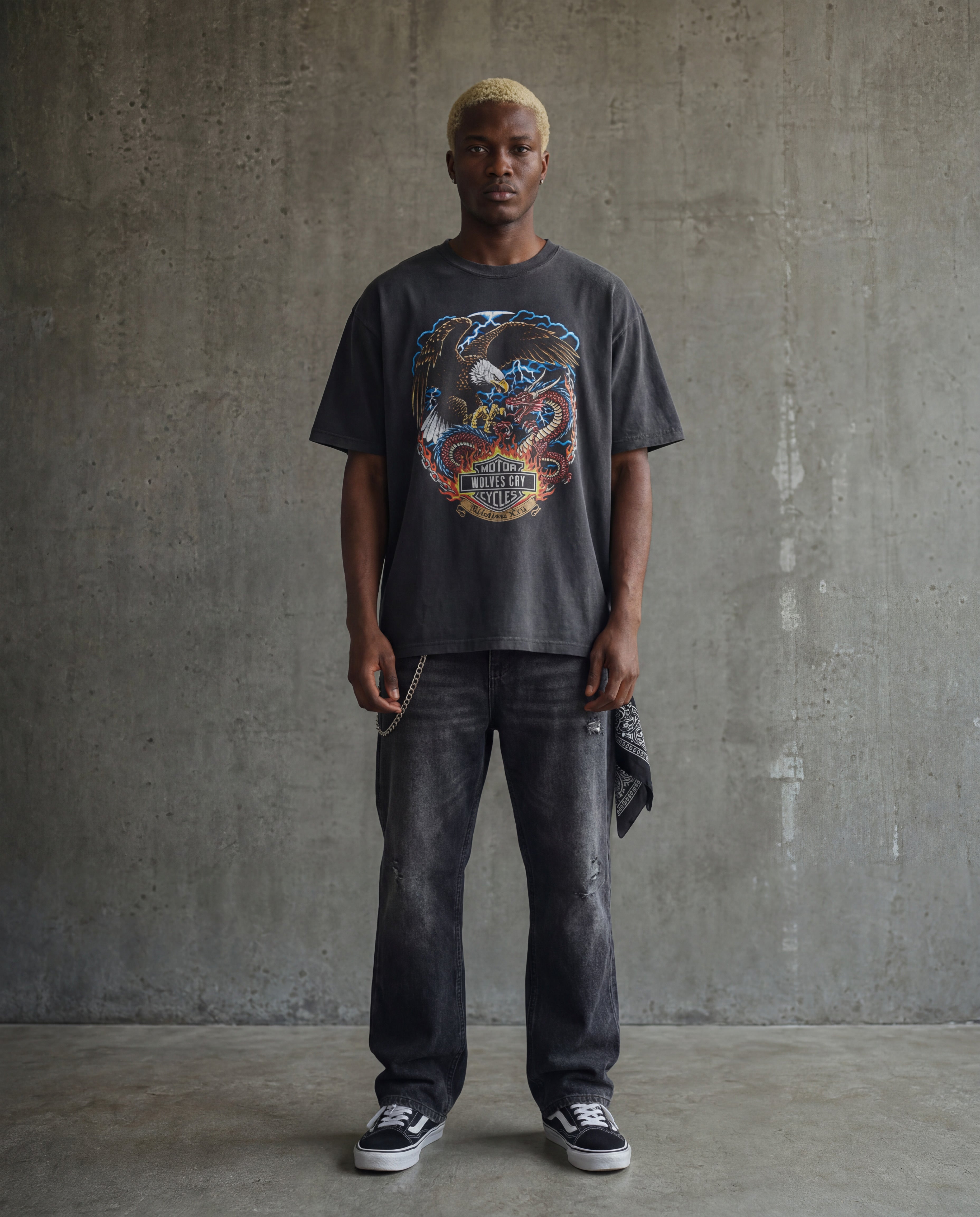 Man wearing a graphic t-shirt and dark jeans against a concrete wall.